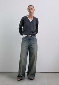 Femeie în picioare cu mâinile la spate, purtând un pulover gri închis cu decolteu în V, un tricou alb pe dedesubt, blugi largi decolorati, curea neagră și sandale cu vârf descoperit.