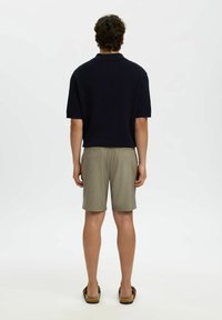Homme de dos portant une chemise noire à manches courtes, un short beige et des sandales marron, sur un fond blanc uni.