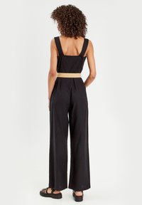 Black linen jumpsuit with wide leg, thick straps, and a woven belt. Features a fitted bodice and a loose fit from waist down. Flat sandals.