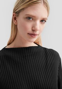 Zwart geribd topje met een brede halslijn, getextureerde stof en lange mouwen, getoond op een model met blond haar en minimale make-up.