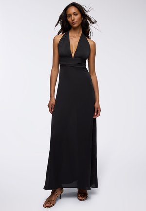 Abito lungo nero con scollatura a V profonda, vita aderente e gonna fluttuante. Tessuto leggero con una consistenza morbida, indossato con scarpe con cinturino.