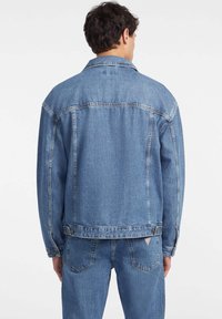 Light blue denim jacket with a classic cut, pointed collar, and two back seams. Features a waist hem with button closure and logo patch.