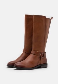 Botas de cuero marrón hasta la rodilla con tacón plano, detalle de correa en el tobillo y acentos metálicos. Textura suave y diseño de puntera redonda.