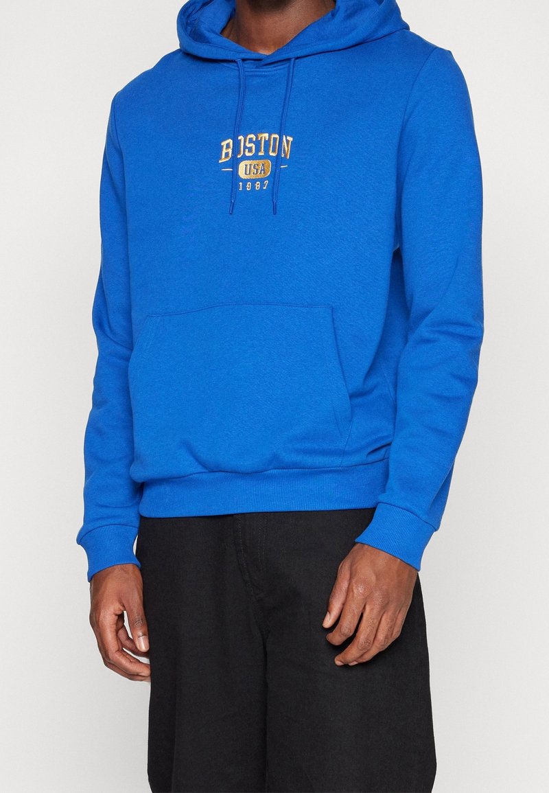 Blå hoodie med en framficka, dragsko på huvan och "BOSTON USA 1902" broderat i gult. Muddar och nederkant är ribbade; tyget är texturerat.