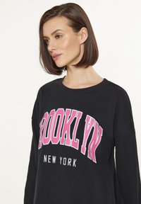 Svart sweatshirt med rosa och vit "BROOKLYN NEW YORK"-tryck. Funktioner inkluderar en rund hals och långa ärmar med en avslappnad passform. Slät tygkvalitet.