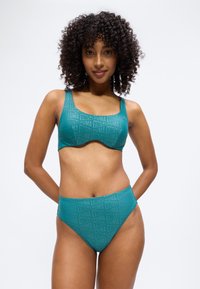 Γυναίκα με σγουρά μαλλιά, που φοράει αθλητικό σουτιέν PUMA σε τιρκουάζ χρώμα και αντίστοιχα σλιπ με ψηλή μέση, στέκεται με τα χέρια πίσω από την πλάτη σε λευκό φόντο.