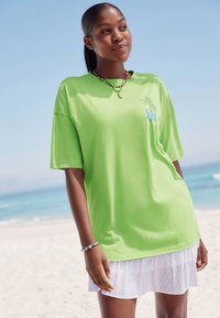 Maglietta verde brillante oversize in tessuto morbido, con una piccola grafica di una palma e la scritta "Soleil." Indossata sopra una gonna bianca a texture.