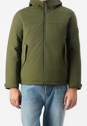 Homme portant une veste à capuche vert olive avec fermeture éclair frontale et poches latérales inclinées, associée à un jean bleu clair.