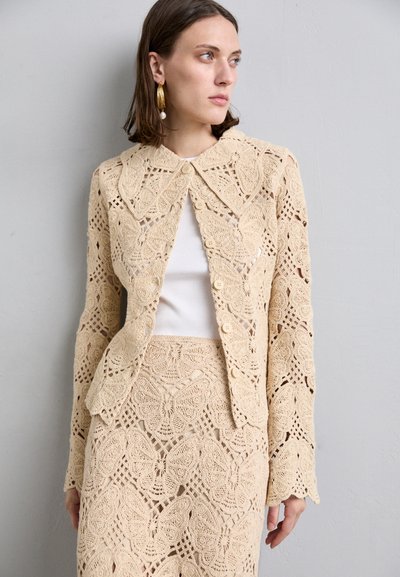 Beige crochet jacket with butterfly patterns, featuring a collared neckline and button closure, paired with a matching skirt. Textured fabric.