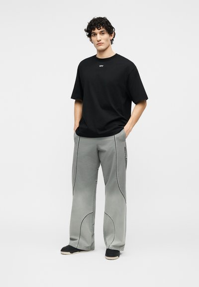 Νεαρός με σγουρά μαλλιά φοράει μαύρο κοντομάνικο μπλουζάκι με το κείμενο "OFF" και χαλαρό γκρι παντελόνι με μαύρες καμπύλες ραφές, στέκεται με τα χέρια στις τσέπες.