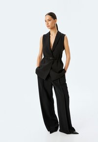 Gilet nero su misura con scollatura a V profonda, vita larga e dettagli con cintura, abbinato a pantaloni neri larghi con una texture liscia.