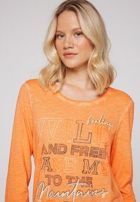 Langärmliges orange T-Shirt aus weichem Stoff, mit Rundhalsausschnitt und Textdruck: "fühlen mich WILD UND FREI, BRING MICH ZU DEN Bergen."