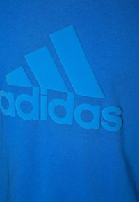 Niebieska bawełniana koszulka z dużym, teksturowanym, ton-sur-ton logo Adidas na środku. Gładka tkanina z okrągłym dekoltem.