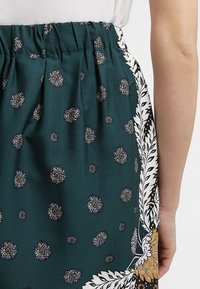 Groene rok met een gestippeld patroon, elastische tailleband en bloemige zijkanten in wit en bruin. Glad stof met een lichte textuur.