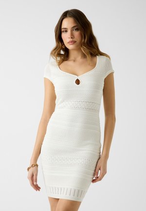 Kobieta ubrana w białą, dzianinową sukienkę z krótkim rękawem i dekoltem w kształcie łezki, stojąca na tle jednolitego, jasnego tła.