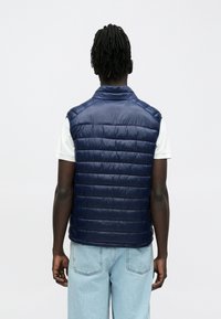 Mann med mørkt flettehår iført en marineblå vattert vest over en hvit kortermet skjorte og lyseblå jeans, vendt bort på hvit bakgrunn.