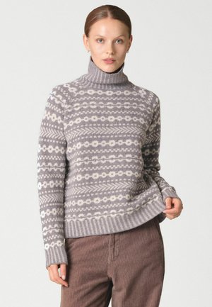 Femme portant un pull à col roulé gris et blanc à motifs et un pantalon en velours côtelé marron, tenant l'ourlet du pull d'une main.