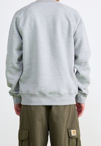 Hellgrau Sweatshirt mit Rundhalsausschnitt und gerippten Bündchen, mit einem Etikett in der unteren rechten Ecke. Getragen mit olivgrünen Cargo-Hosen.