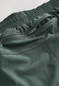 Pantalon à taille élastique vert foncé avec une poche zippée fermée et des coutures visibles sur une surface de tissu lisse.