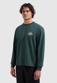 Man met krullend haar, gekleed in een donkergroene longsleeve shirt met een klein wit logo op de linkerborst en zwarte broek, staande tegen een grijze achtergrond.