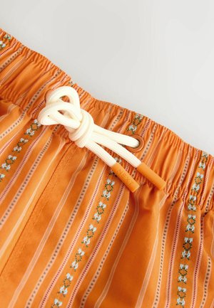 Orange gestreifter Stoff mit Blumenmustern, elastischem Bund und weißem Kordelzug mit orangefarbenen Endkappen, auf denen "NEXT" eingraviert ist, zu einer Schleife gebunden.