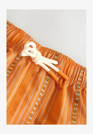 Pomarańczowa tkanina w paski z kwiatowymi wzorami, elastyczny pas i biały sznurek ze sznurówkami zakończonymi pomarańczowymi nasadkami z wygrawerowanym napisem "NEXT", zawiązany na kokardę.