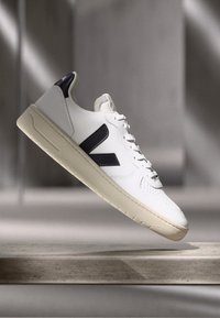 White leather sneaker with black accents, textured side panels, and a thick, cream-colored sole, featuring a logo on the outer side.