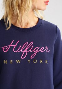 Sweat-shirt bleu marine avec un col rond, orné du texte brodé en rose "Hilfiger" et de "NEW YORK" en dessous en or. Texture de tissu douce.
