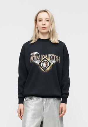 Femme blonde portant un sweat-shirt noir avec l'inscription « VOM RUTCH » et un emblème ailé, assorti à un pantalon métallisé argenté, se tenant devant un fond blanc.