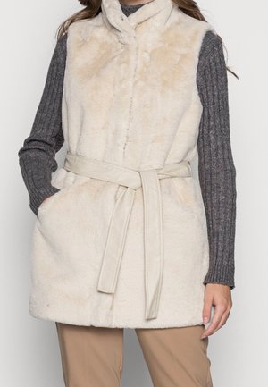 Veste sans manches en fausse fourrure crème avec ceinture, portée sur un pull gris côtelé à manches longues et un pantalon beige, mains dans les poches.