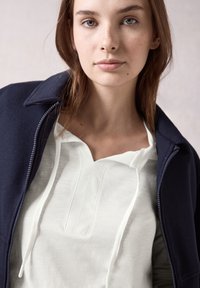 Marine blauwe zip-up jas over een witte langmouwtop met een gestructureerde afwerking en een keyhole halslijn. Natuurlijke haren, minimale make-up.