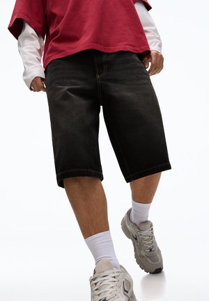 Osoba nosi čierne džínové šortky po kolená, biele ponožky, béžové tenisky, dlhý biely rukáv pod červeným krátkym rukávom.