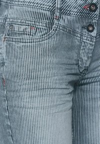 Randiga jeans i ljusblå denim med smala vertikala vita ränder, utrustade med två knappar och röda sömnadsdetaljer, texturerade med en slät finish.