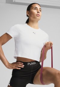 Hvid cropped atletisk shirt med korte ærmer og grå Puma-logo, parret med sorte shorts med en hvid Puma-linning, mens der holdes en rød modstandsbånd.