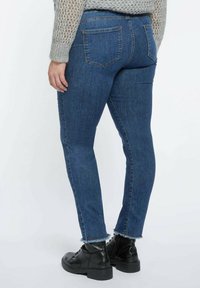 Persona con pantalones vaqueros de mezclilla azules con dobladillos deshilachados, botas negras con cordones y un suéter gris de punto, de pie contra un fondo blanco.