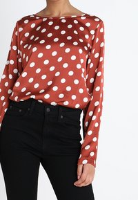 Braun glänzendes Langarmoberteil mit großen weißen Punkten. Kombiniert mit hoch geschnittenen schwarzen Jeans. Glatte Textur mit lockerer Passform.