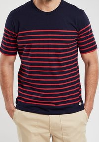 Hombre vestido con una camiseta azul marino con rayas horizontales rojas y pantalones beige, con las manos casualmente en los bolsillos.
