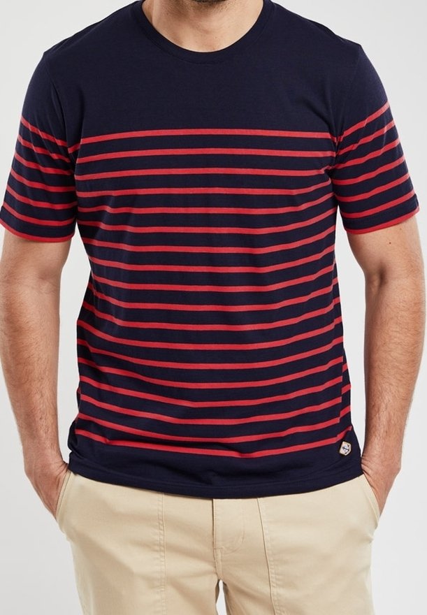 Hombre vestido con una camiseta azul marino con rayas horizontales rojas y pantalones beige, con las manos casualmente en los bolsillos.
