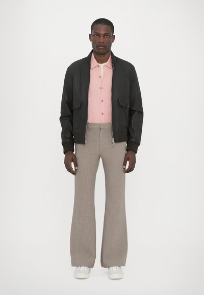 Chaqueta bomber negra con dos bolsillos frontales, sobre una camisa de botones rosa claro, combinada con pantalones de campana beige y zapatillas blancas.