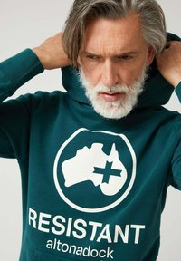 Sudadera con capucha color verde azulado hecha de tela suave que presenta un mapa de Australia y un signo de más en blanco, con el texto "RESISTENTE" debajo en una fuente negrita y contrastante.