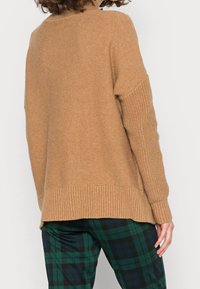 Sweter w kolorze camel, dzianinowy, z prążkowanym dołem i mankietami. Ma luźny krój i teksturowany panel na rękawach. Połączony z spodniami w kratę.