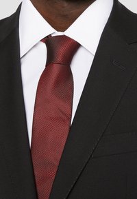 Zwarte blazer met scherpe revers, wit overhemden en een gestructureerde bordeauxrode stropdas met een fijn patroon.