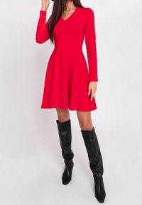 Rotes langärmliges Kleid mit V-Ausschnitt und enganliegenden Oberteil, das sich in einen ausgestellten Rock entfaltet. Kombiniert mit knielangen schwarzen Lederstiefeln.