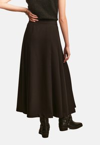 Langer schwarzer Rock aus einem glatten, fließenden Stoff. Mit einem figurbetonten Bund und ausladendem Saum, kombiniert mit schwarzen Stiefeletten.