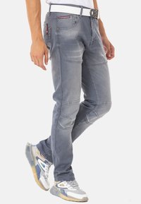 Graue Jeans aus Denim mit schmalem Schnitt, ausgestattet mit Knietafeln, dezenten Stickdetails und einem weißen Gürtel. Stylische Taschen setzen funktionale Akzente.