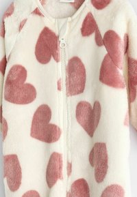 Rosa und weiße Fleece-Einteiler mit einem Frontreißverschluss, der ein Herzmuster in unterschiedlichen Größen und einer weichen Textur aufweist. Mit einem gerippten Kragen akzentuiert.
