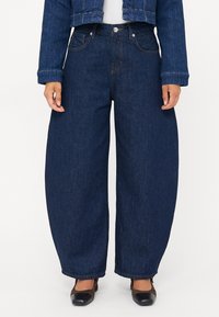 Moteris vilki tamsiai mėlynas plačias kelnes, trumpą džinsinį švarkelį, baltą marškinėlį ir juodai rudos uždaros nosies aukštakulnius batus ant balto fono.