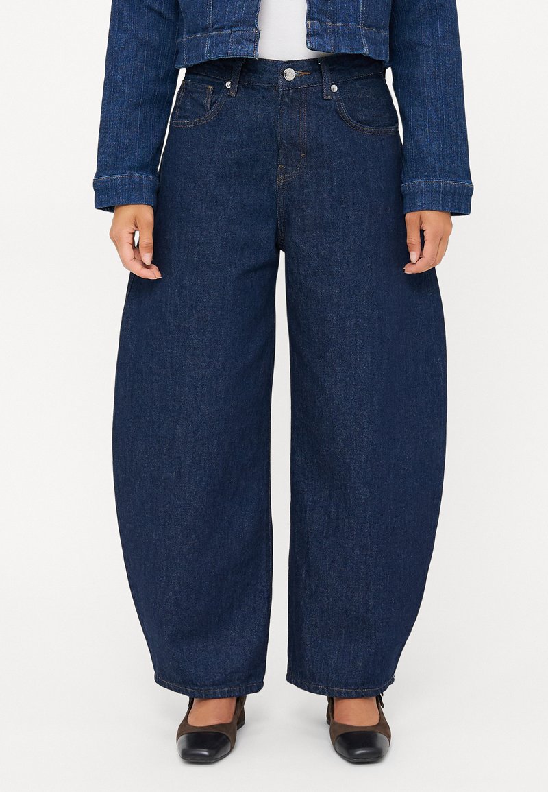 Moteris vilki tamsiai mėlynas plačias kelnes, trumpą džinsinį švarkelį, baltą marškinėlį ir juodai rudos uždaros nosies aukštakulnius batus ant balto fono.