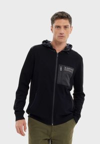 Felpa nera con zip, dal design aderente, caratterizzata da una grande tasca frontale testurizzata, cappuccio regolabile e polsini a costine. Tessuto morbido.