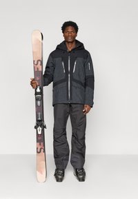 Echipamentul de schi include o jachetă neagră cu glugă și modele texturate, pantaloni negri și schiuri cu accente maro închis și deschis. Încălțămintea este neagră.